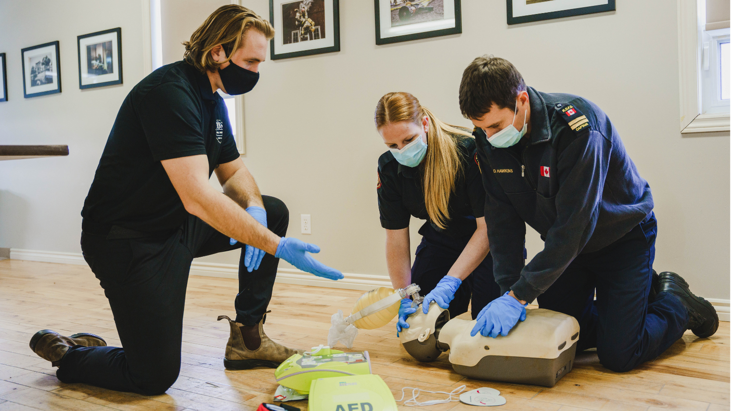 Three people practicing CPR and using an AED in a room with framed pictures on the wall.